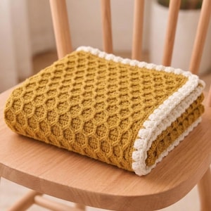 May include: A folded mustard yellow crochet blanket featuring a textured honeycomb stitch pattern throughout the center. The blanket is finished with a thick, white, scalloped crochet border that frames the entire rectangular piece.