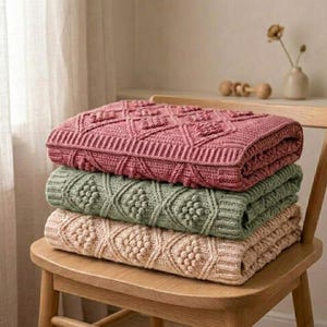 Peut inclure: Une pile de trois couvertures en crochet pliées dans des tons rose poudré, vert sauge et crème. Chaque couverture présente un motif en losanges texturé avec des points popcorn en relief et une bordure côtelée, posée sur une chaise en bois.