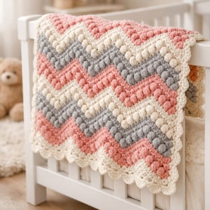 May include: A crochet blanket featuring a chevron pattern with alternating stripes of cream, soft pink, and grey yarn. The design incorporates raised bobble stitches along each chevron row, finished with a scalloped edge border in cream.