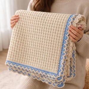 May include: A cream-colored waffle-knit blanket featuring a decorative border with a light blue yarn trim and a scalloped edge pattern. The blanket has a textured grid design throughout the center and is folded to display the detailed crochet edging.