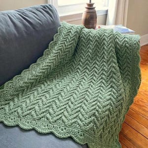 May include: A sage green crochet blanket with a chevron pattern. The blanket is draped over a blue sofa, highlighting its detailed design and scalloped edge. The blanket's colour and pattern create a cosy, inviting aesthetic, perfect for home decor.