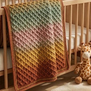 May include: A crochet blanket featuring a textured waffle stitch pattern with a gradient colour transition from sage green to soft pink, golden yellow, and deep burnt orange, finished with a solid border draped over a wooden crib railing.