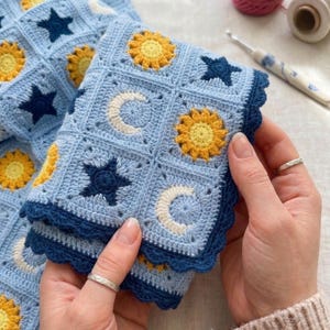 Könnte beinhalten: Hellblaue Häkeldecke mit quadratischen Mustern, die gelbe Sonnenblumen, dunkelblaue Sterne und weiße Halbmonde zeigen. Die Decke hat einen dunkelblauen, gewellten Rand. Gehalten von Händen.