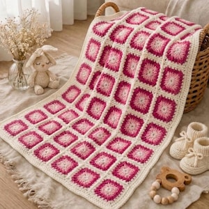 May include: A cream and pink crochet baby blanket with a square pattern, draped over a wicker basket. The image also shows a crochet bunny, baby booties, and a wooden teether. The blanket is likely 100cm x 100cm.