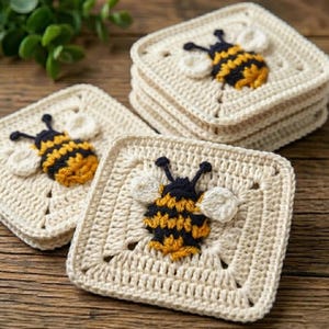 May include: A set of square crochet coasters in a cream color, each featuring a central three-dimensional bumblebee motif. The bees are crafted with black and yellow yarn stripes and white wings, centered on the textured, open-stitch square base.