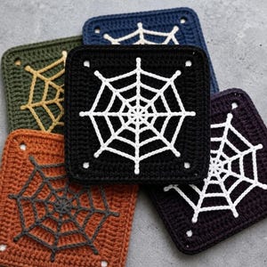 May include: A collection of five square crochet coasters featuring spider web patterns. The coasters are in black, dark blue, olive green, orange, and dark purple, each with a contrasting white or dark gray web design stitched onto the center.