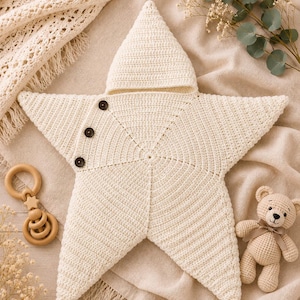 May include: A cream-colored star-shaped crochet swaddle blanket featuring a hood and three dark brown buttons along one side. The item is accompanied by a small wooden ring rattle and a miniature crocheted teddy bear with a brown ribbon.