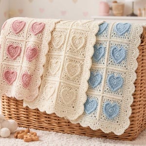 Op de afbeelding: Drie gehaakte babydekens met hartontwerpen in roze, crème en blauw worden in een rieten mand getoond. Elke deken heeft een geschulpte rand en een rasterpatroon van hartvormige applicaties, perfect voor een kinderkamer.