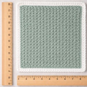 May include: A square crochet washcloth featuring a sage green center with a textured stitch pattern and a solid white border. The washcloth is positioned next to wooden rulers, showing it measures approximately 8 inches in width and height.