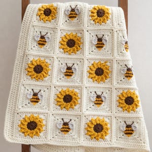 May include: A handmade crochet blanket featuring a repeating grid pattern of off-white squares. Each square contains either a textured, three-dimensional yellow sunflower with a dark brown center or a small bee with yellow and black stripes.