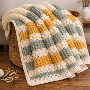 May include: A handmade crochet blanket with alternating stripes of cream, mustard yellow, and muted green. The blanket features a textured waffle stitch and bobble details, folded over a wicker basket. A ball of yarn and scissors are on the wooden surface.