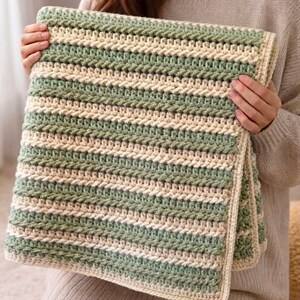 May include: A rectangular crochet blanket featuring alternating horizontal stripes of sage green and cream yarn. The textile displays a consistent textured stitch pattern throughout, finished with a simple cream-colored border along all four edges.
