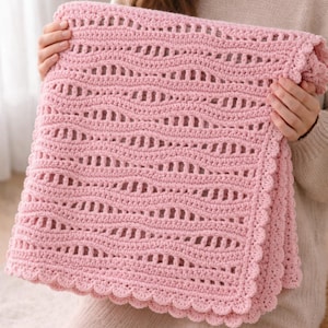 May include: A soft pink crochet blanket with a scalloped edge. The blanket features a repeating wave pattern throughout the design. The blanket is folded and held up, showcasing the texture and color.