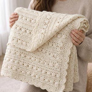 Puede incluir: Manta de ganchillo de color crema con un patrón floral detallado. La manta tiene un borde festoneado y está doblada, mostrando el intrincado trabajo de puntadas. La manta suave y texturizada se sostiene, destacando su calidad hecha a mano.