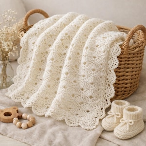May include: A cream-colored, crocheted baby blanket with a scalloped edge is draped over a wicker basket. Matching crocheted baby booties with bow details and a wooden teething ring are also present, creating a cozy, handmade theme.