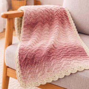 May include: A handmade, wave-patterned blanket in shades of pink and cream, draped over a wooden chair. The blanket features a scalloped edge and a gradient color scheme, transitioning from a deep rose to a soft cream color.