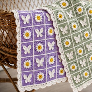 Könnte beinhalten: Eine handgefertigte Häkeldecke mit einem Gitter aus quadratischen Feldern in Lavendel und Salbeigrün. Jedes Quadrat enthält entweder ein weißes Gänseblümchen mit gelber Mitte oder eine weiße Schmetterlingsapplikation. Weißer Häkelrand.