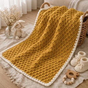 Può includere: Coperta per bambini giallo senape con motivo a nido d'ape e bordo bianco. La coperta è piegata su un cestino di vimini. Un coniglietto all'uncinetto, scarpine per bambini e un anello per la dentizione in legno sono anche nell'immagine.