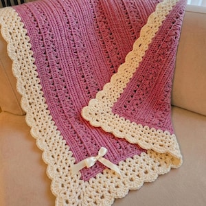 May include: A handmade crochet blanket in a soft pink hue, featuring a detailed stitch pattern. The blanket is trimmed with a cream-colored, scalloped border. A cream-colored ribbon is tied in a bow at the corner.