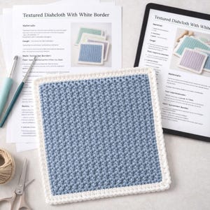 May include: A square crochet dishcloth with a textured blue center and a white border, placed on top of printed instruction sheets titled Textured Dishcloth With White Border, alongside two crochet hooks, a ball of yarn, and metal scissors.