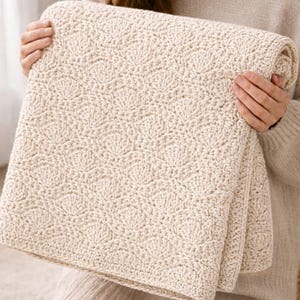 May include: A folded, cream-colored blanket with a textured, shell-like pattern. The blanket is made of a soft, knitted material. The blanket is held by a person wearing a cream-colored sweater.