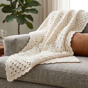 Peut inclure: Plaid de crochet crème drapé sur un canapé gris. La couverture présente un motif de tissage ouvert texturé avec un bord festonné. Un coussin marron est visible sur le canapé. Une plante en pot est en arrière-plan.
