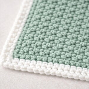 May include: A square crochet textile featuring a sage green center with a textured moss stitch pattern. The piece is finished with a clean, white crocheted border that frames the edges, creating a distinct contrast between the two colors.