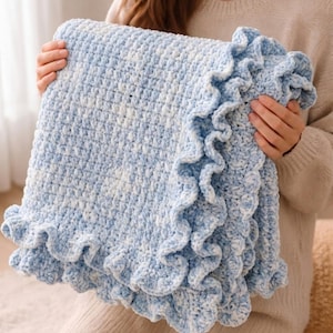 May include: A soft, chunky crochet blanket featuring a variegated pattern of light blue and white yarn. The rectangular blanket is constructed with a dense, textured stitch and is finished with a decorative, ruffled border along the edges.