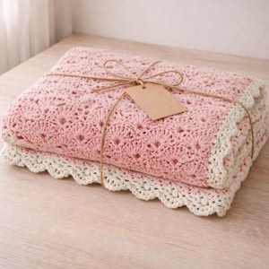 May include: A folded pink crochet blanket with a cream-colored scalloped edge, tied with twine and a blank tag. The blanket is made of soft yarn and is perfect for a baby or as a decorative throw.