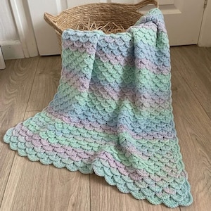 Könnte beinhalten: Handgefertigte Häkeldecke mit einem Fischschuppenmuster. Die Decke hat einen Farbverlauf von Hellblau, Mintgrün und Lavendel. Sie liegt auf einem Holzboden und ist über einem geflochtenen Korb drapiert.