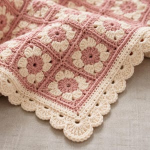 May include: A crochet blanket featuring a repeating pattern of square motifs with dusty rose centers and cream-colored petals. The blanket is finished with a decorative scalloped edge in cream yarn, showcasing intricate hand-stitched texture.