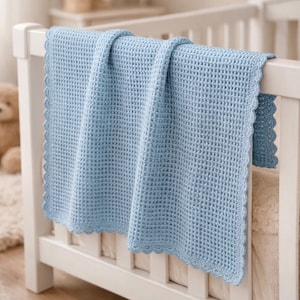 May include: A light blue crochet blanket featuring a textured waffle stitch pattern and a decorative scalloped edge. The blanket is draped over the side of a white wooden crib, showcasing the uniform square grid design and the soft yarn texture.