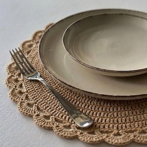 Op de afbeelding: Een roestvrijstalen vork ligt op twee gestapelde, gebroken witte keramische borden met donkerbruine randen, geplaatst op een ronde, beige gehaakte placemat met een geschulpte rand en een dicht, gestructureerd steekpatroon in het ontwerp.