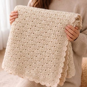 May include: A cream-colored, crocheted blanket with a scalloped edge. The blanket features a detailed openwork pattern. The soft, neutral color and intricate design suggest a handmade item, perfect for home decor or as a cozy accessory.
