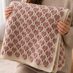 May include: A crocheted blanket featuring a heart pattern in shades of pink and cream. The blanket is folded, showcasing the intricate stitchwork and texture. The heart design is repeated across the surface, creating a cozy and decorative textile.