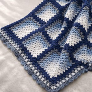 May include: A handcrafted crochet blanket in various shades of blue, white, and grey. The blanket has a square pattern and a scalloped edge. The colour scheme creates a calming and cosy aesthetic, suitable for home decor.