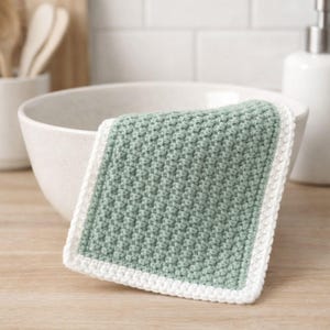 May include: A square crochet washcloth featuring a textured sage green center panel and a solid white border. The fabric displays a consistent, dense stitch pattern throughout the main body, framed by a clean, simple white edge finish.