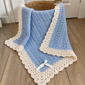 May include: A light blue crochet blanket featuring a textured stitch pattern and a wide, cream-coloured scalloped border. The blanket is accented with a small, white satin ribbon bow tied at the bottom edge, creating a soft and detailed appearance.