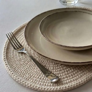 Op de afbeelding: Een ronde, crèmekleurige gehaakte placemat met daarop twee gestapelde, gebroken witte keramische borden met donkere, rustieke randen. Een gepolijste roestvrijstalen vork met vier tanden ligt aan de linkerkant van de placemat en overlapt het onderste bord.