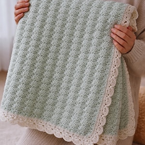 May include: A sage green crocheted blanket featuring a textured shell stitch pattern throughout the main body. The blanket is finished with a wide, cream-colored scalloped crochet border that frames the edges with a delicate, decorative design.