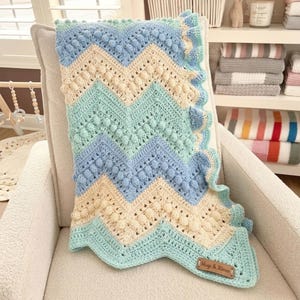 May include: A crochet blanket featuring a chevron pattern in light blue, mint green, and cream. The design includes raised bobble stitches along the chevron rows and a ruffled edge. A brown rectangular leather tag is attached, reading Hugs & Kisses.