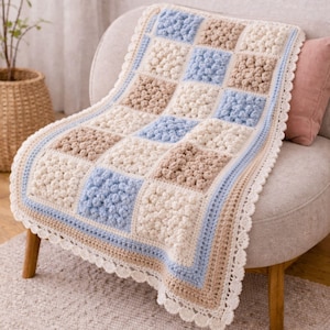 May include: A handmade crochet blanket in a patchwork design, featuring squares of cream, light blue, and tan. The blanket has a scalloped edge and is draped over a chair. The blanket is a cozy home decor item.