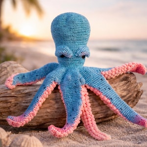 May include: A handmade crochet octopus in shades of blue and pink. The octopus is sitting on a piece of driftwood on a sandy beach. The tentacles are blue with pink tips. The background shows a sunset over the ocean.