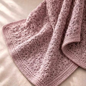 Könnte beinhalten: Eine staubrosa gehäkelte Decke mit floralem Muster. Die Decke hat ein detailliertes Stichdesign mit einem durchgehenden Rand. Die weiche, strukturierte Decke ist gefaltet und zeigt die komplizierte Handwerkskunst und das Design.