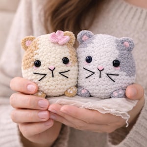 May include: Two handmade crochet cat toys. One is beige and tan with a pink flower, the other is gray and white. Both have black eyes, pink accents, and black whiskers. The toys are held in cupped hands.