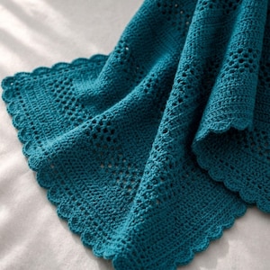 May include: A teal crocheted blanket with a scalloped edge. The blanket has an openwork design and a textured pattern. The soft, handmade blanket is folded, displaying its intricate details and craftsmanship.