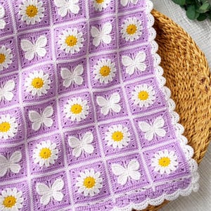 May include: A handmade crochet blanket featuring a repeating pattern of white butterflies and daisies with yellow centers on a lavender background. The blanket has a white scalloped edge and is displayed on a woven basket.
