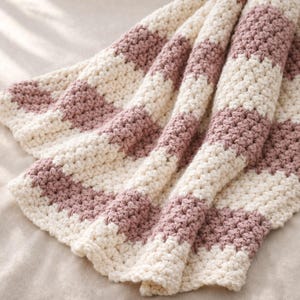 May include: A hand-crocheted blanket featuring alternating horizontal stripes of cream and dusty rose yarn. The textile displays a consistent, textured stitch pattern throughout, creating a soft, folded appearance with visible depth and dimension.