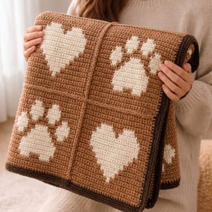 May include: A square crochet blanket featuring a grid pattern with alternating cream-colored heart and paw print motifs on a medium brown background, finished with a dark brown border. The textile displays a consistent stitch texture throughout.