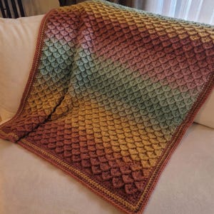 May include: A handmade crochet blanket in a gradient of colors, transitioning from a deep reddish-brown to yellow and green. The blanket features a textured, scale-like pattern and a solid border.
